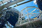 From the bottom right-hand corner of the image, reaching across half the image towards the left-hand corner, spans the white beams of the main structure. Above the viewer is a platform of grey scaffolding. Underneath on the left of the main structure and on the right, are the unfinished dark grey panels of the dome, with a skeleton structure of scaffolding underneath. Blue sky spans above the open construction site, with a small wispy cloud above the right side of the dome.