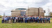 This photo shows a large group of ESO staff members and representatives from other industries and institutions standing outside the rounded, curvy ESO Supernova building. This large number of smartly-dressed individuals are standing on green grass just in front of this large, metallic-brown structure with its huge blue windows. Everyone in the image is wearing a blue lanyard, and a smile.